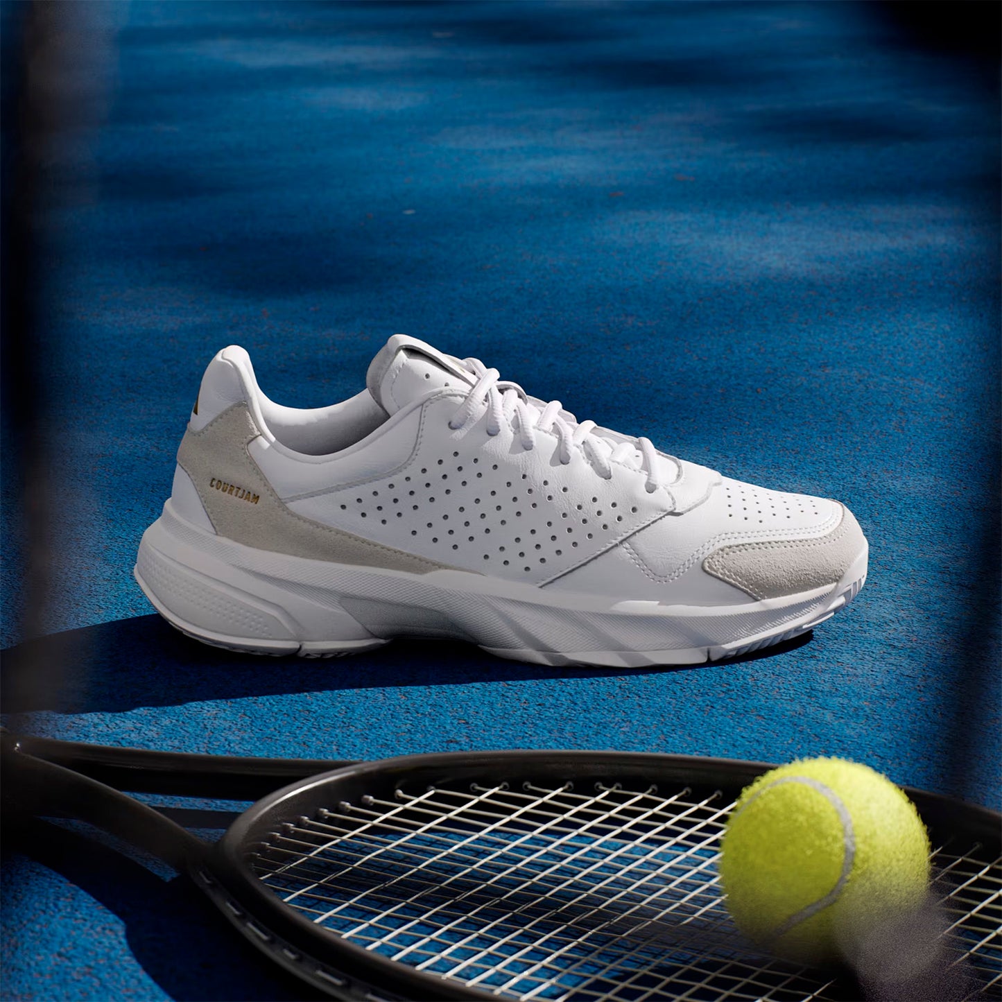 White Adidas Unisex Courtjam Control 3 Premium shoe on a blue tennis court with a racket and yellow ball.
