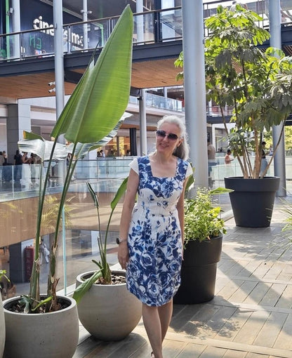 Woman in a blue floral dress standing among potted plants in an outdoor setting.