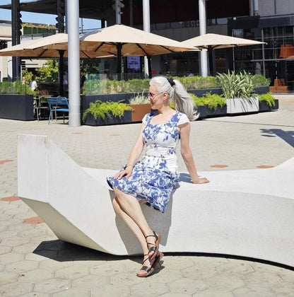 Woman in a blue floral dress sitting on a modern outdoor bench with plants and umbrellas in the background.